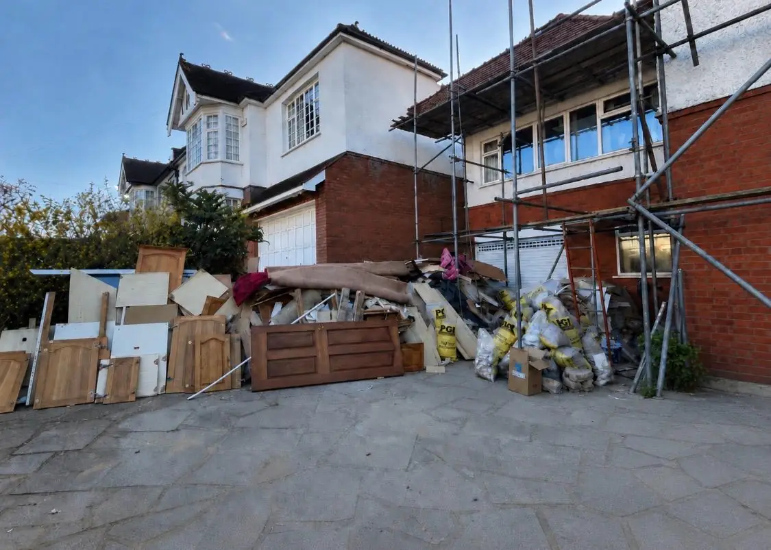 A house driveway part way through a clearance, internal doors and sheets of ply stacked on edge, a rolled carpet and filled rubble bags across the flagstones, scaffolding on the house behind